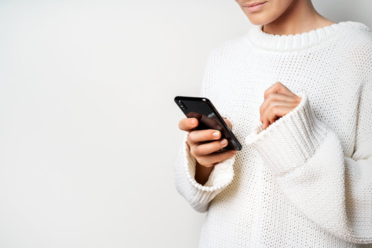 A young woman in a white sweater holds a smartphone in her hands, chooses something looking at the screen.