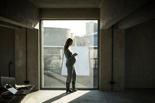 Pregnant businesswoman standing at office window, using smartphone - Powered by Adobe