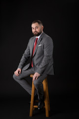 portrait of a bearded fashion man wearing at suit while seated on a high chair and looking into the camera. on a black background