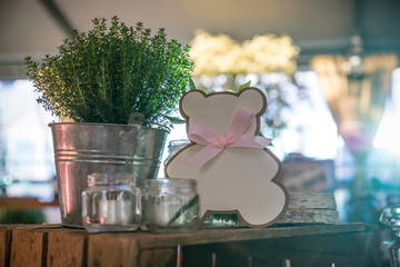 Garden composition with potted thyme plant, white roses, candles in a glass jar and a pink cardboard bear, on a wooden box in a rustic setting