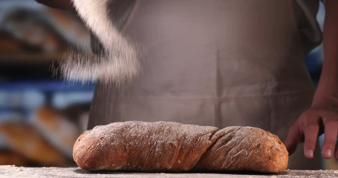 In the bakery, a baker after baking fresh bread sprinkles sugar powder (flour) on bread (flour pastry) with a powder colander (with hands). Concept of: Fresh bread, Home-made, The hands, Slow Motion.