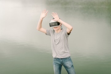 Young adolescent playing a virtual reality simulation with vr glasses and doing hand gestures outdoors near a lake