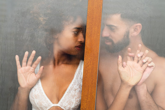 Handsome Hispanic guy touching and kissing seductive African American woman in lace bra while standing behind wet window at home