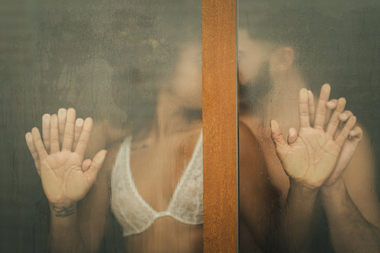 Handsome Hispanic guy touching and kissing seductive African American woman in lace bra while standing behind wet window at home