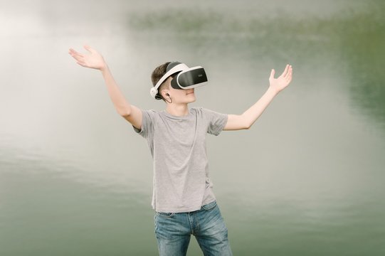 Young adolescent playing a virtual reality simulation with vr glasses and doing hand gestures outdoors near a lake