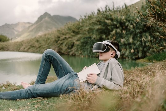 Young adolescent with virtual reality glasses laying on grass outdoors near a lake with a book