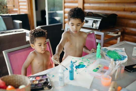 Two shirtless African American boys using bright paint to make abstract pictures on table at home
