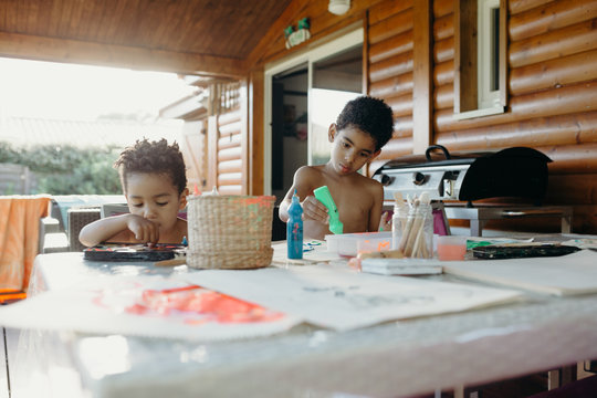 Two shirtless African American boys using bright paint to make abstract pictures on table at home