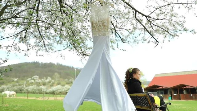 Wedding long dress hanged on a wooden hanger loosely on a tree fluttering in the wind during a sunny day before the ceremony