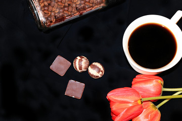 A cup of coffee, chocolate and tulips on a black background close-up, selective focus. Elegant design of your evening