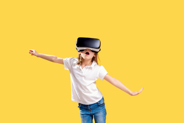 Girl 7 y.o in formal outfit wearing VR glasses putting hands out in excitement isolated on yellow background. Child using a gaming gadget for virtual reality. Virtual technology.