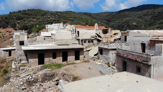 Broken houses at abandoned village, dark gaps of empty doors and windows, destroyed walls and debris around. Red roof of unbroken or rebuild church seen at back, green hills on background