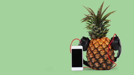 fresh pineapple with black headphones and smartphone with isolated black screen against green background.