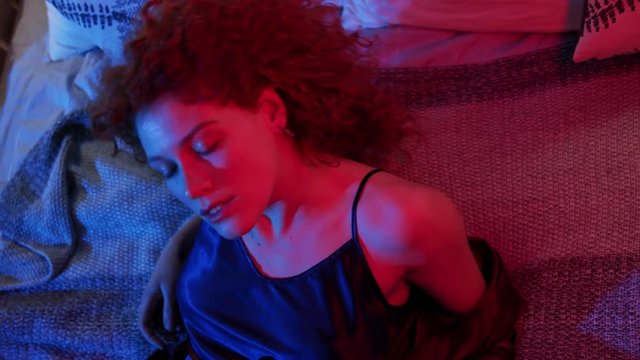 Young woman playfully looks straight to camera and lies on bed. Romantic date, seductive outfit, provoking. Playful mood, positive emotions. Top close up view. Female portrait in neon light