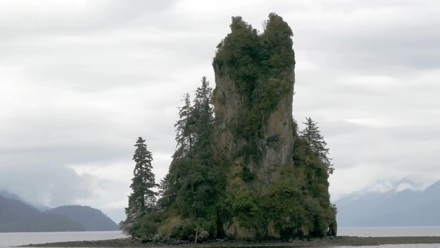 A close shot of New Eddystone Rock, a massive rock formation that sits on an isle in Alaska. The ocean and mountains are visible in the distance.