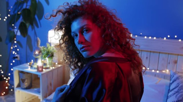 Pretty young Caucasian curly woman playfully covering her naked shoulders with black nightgown and seductively looks straight to camera in neon light. Sexy brunette lover in underwear lying on bed