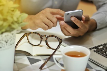 man typing smartphone on desk office with coffee cup, glasses and small tree