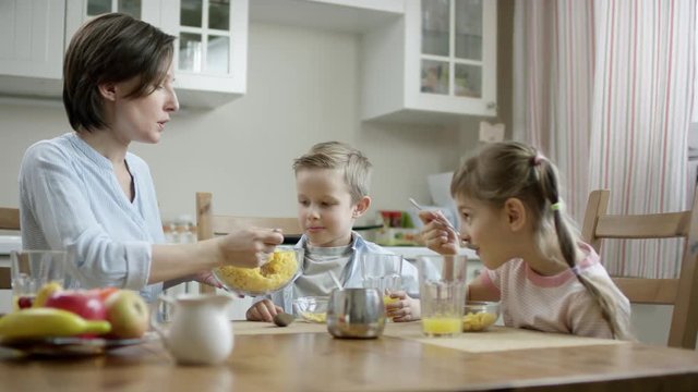 mother put cornflakes from bowl on the plate with a spoon to her son while daughter eating