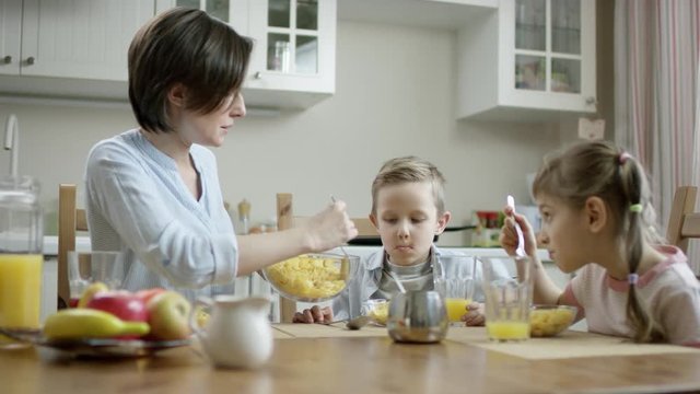 mother communicate during breakfast with her children and put cornflakes from bowl on the plate with a spoon to her son