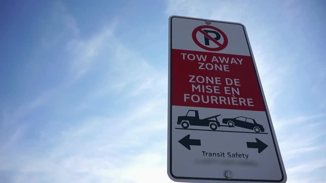 Panning right to no parking sign on a clear blue sky day. Panning right to tow away zone transit safety sign French. A bilingual notice sign, says Tow away zone.