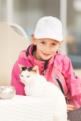 Young blond girl, primary school age, wearing sun cap, caressing cat lying on sunbed