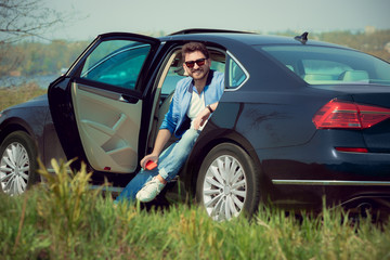 Handsome smiling man in jeans, jacket and sunglasses sitting in his car with opened doors on the river's side. Preparing for weekend' trip or journey. Concept of summertime, resort, chilling.