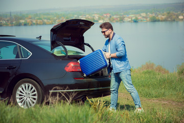Handsome smiling man in jeans, jacket and sunglasses going to vacations, loading his suitcase in car trunk on the river's side. Preparing for weekend' trip. Concept of summertime, resort, chilling.
