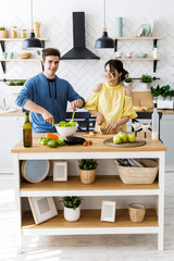 Beautiful couple in kitchen. Handsome man is preparing a salad while his girlfriend is cutting bread, both are smiling and have fun time