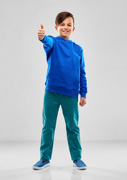 childhood, gesture and people concept - portrait of smiling boy in blue hoodie showing thumbs up over grey background