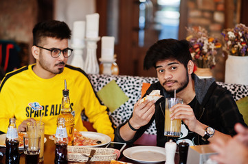 Asian friends guys eating pizza during party at pizzeria. Happy indian people having fun together, eating italian food and sitting on couch.