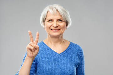 gesture and old people concept - portrait of smiling senior woman in blue sweater showing peace over grey background