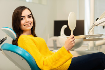 Young happy woman client looking at the mirror with toothy smile at the dental office