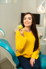 Happy girl sitting in dental chair and showing fresh apples after successful dental treatment