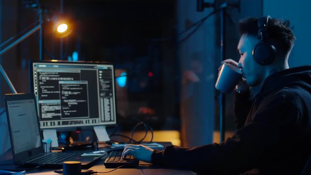 cybercrime, hacking and technology concept - asian male hacker in headphones with coding on laptop computer screen wiretapping or making cyber attack and drinking coffee in dark room at night