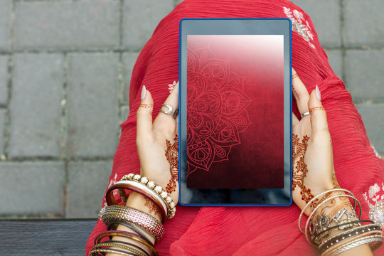 Beautiful woman wear traditional Muslim Arabic Indian wedding red pink sari dress hands with henna tattoo mehndi pattern jewelry and bracelets hold tablet. Summer culture festival celebration concept.