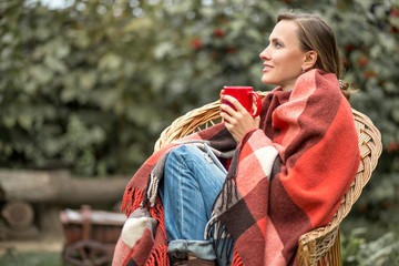Beautiful girl resting and drinknig coffee sitting in autumn garden in a chair wrapped in a plaid...