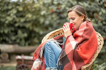 Beautiful girl resting and drinknig coffee sitting in autumn garden in a chair wrapped in a plaid woolen blanket