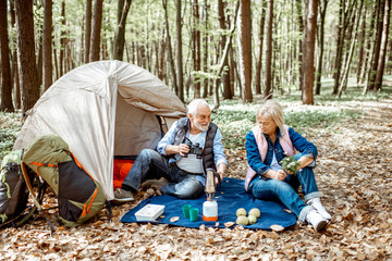 Senior couple sitting together at the campsite with tent and backpacks, enjoying nature with binoculars in the forest