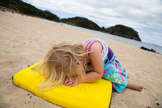 Pre school girl having a meltdown at the beach because she doesn't want to go home. Beautiful day, white sand, clear blue sea. Girl lying on a yellow board with hair covering face. Dramatic angle