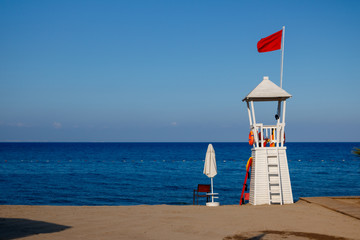 Empty lifeguard post with a red flag at a white, beach under a clear sky.