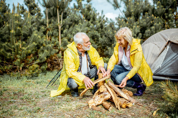 Senior couple in yellow raincoats making fireplace at the campsite near the tent in the woods