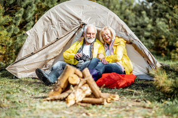 Senior couple in yellow raincoats sitting together near the tent at the campsite with fireplace in the woods