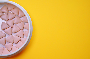 Pink triangular unusual pills in a white round box on a yellow background with copyspace, top view, macro