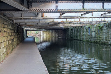 view under an old low steel girder bridge crossing the leeds to liverpool canal near armley with stone wall and a narrow footpath alongside the water