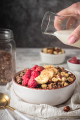 healthy breakfast granola in a plate with nuts, banana and raspberries, milk is poured from a bottle, copy space