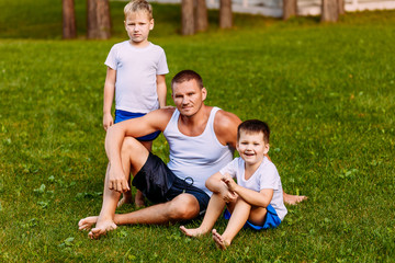 Naklejka na ściany i meble father and two children sit on the green lawn in the summer. A man and two young sons of a preschooler in the summer