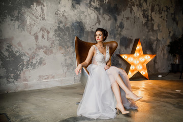 Young beautiful wedding bride in a beautiful long dress sitting in a chair in the apartment in the...