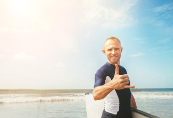 Young man Surfer portrait showing surfer's famous Shaka sign gesture in camera when he comming with long surf board to waves. Active holidays spending concept.