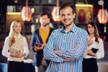 Young attractive arab businessman standing at restaurant with arms crossed. In background his successful team posing.