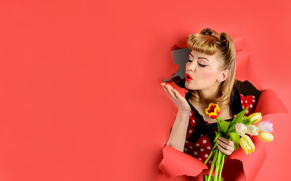 Happy woman through paper sends air kiss. Pin up woman looking through paper blowing kiss with hand. Sexy woman holds flowers. Bouquet of tulips. Breaking paper. retro hairstyle. Spring sale. Discount